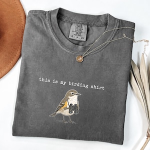 May include: A charcoal gray crew neck t-shirt featuring a graphic of a small bird holding black binoculars. The text "this is my birding shirt" is printed in white above the bird. The shirt has a Comfort Colors brand label at the inner neckline.