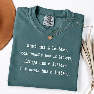 May include: A teal crewneck t-shirt featuring white text that reads: what has 4 letters, occasionally has 12 letters, always has 6 letters, but never has 5 letters. The shirt has a Comfort Colors brand label inside the collar and a gold-toned necklace.