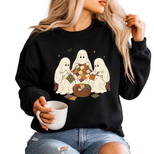 May include: A black long-sleeve sweatshirt featuring a graphic print of three white ghosts sitting together and quilting. The ghosts are working with colorful patterned fabric squares and a wicker basket filled with sewing supplies and fabric scraps.
