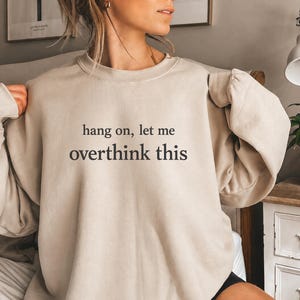 May include: A beige crewneck sweatshirt featuring the black text "hang on, let me overthink this" printed in a serif font across the chest. The garment has long sleeves, a ribbed neckline, and a relaxed fit, providing a simple and clean design.