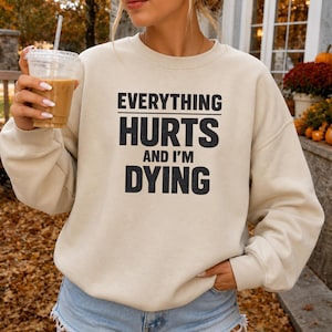 May include: A beige crewneck sweatshirt featuring the black printed text "EVERYTHING HURTS AND I'M DYING" centered on the chest. The garment has long sleeves, a ribbed neckline, and ribbed cuffs, providing a standard fit for casual wear.