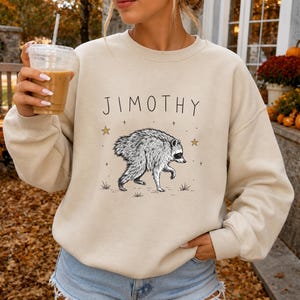 May include: A beige crewneck sweatshirt featuring a black line art illustration of a raccoon walking. Above the raccoon, the text JIMOTHY is printed in a simple black font. Small gold stars and tiny black crosses are scattered around the raccoon.