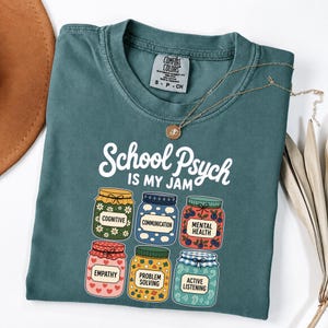 May include: A teal crewneck t-shirt featuring the white text School Psych Is My Jam above six colorful jars labeled Cognitive, Communication, Mental Health, Empathy, Problem Solving, and Active Listening, with a gold necklace resting on the collar.