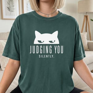 May include: Dark green t-shirt with a white cat graphic and the text "JUDGING YOU SILENTLY." The shirt has short sleeves and a relaxed fit. The cat graphic features a simple, minimalist design.