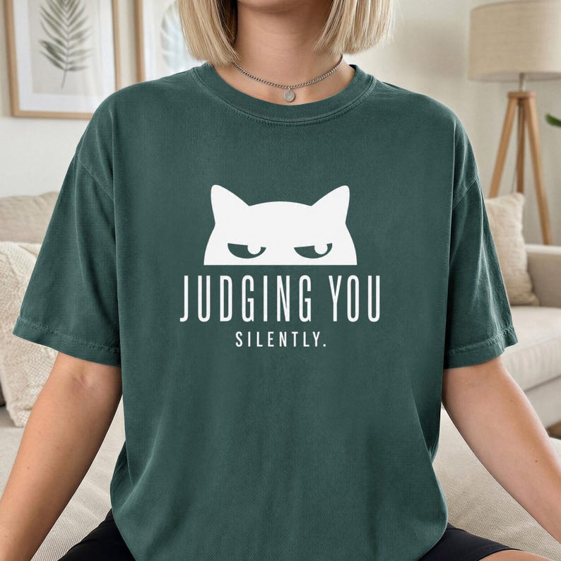 May include: Dark green t-shirt with a white cat graphic and the text "JUDGING YOU SILENTLY." The shirt has short sleeves and a relaxed fit. The cat graphic features a simple, minimalist design.