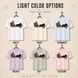 May include: Six short-sleeve t-shirts in white, ivory, chambray, orchid, bay, and blossom colors. Each shirt features a graphic of a brown tabby cat sitting in a chair reading and a black cat lying on a couch. Text includes LIGHT COLOR OPTIONS.