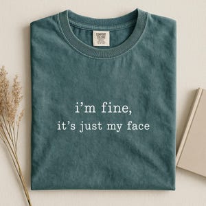 以下が含まれることがあります： 胸の中央に白い文字で「i'm fine, it's just my face」とプリントされた、ダークティールカラーのクルーネックTシャツ。リブ編みの襟と、首の内側にComfort Colorsのブランドラベルが付いた、無地のデザインのTシャツです。