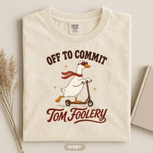 Könnte beinhalten: Ein elfenbeinfarbenes T-Shirt mit einer Grafik einer weißen Gans, die eine braune Fliegerbrille und einen roten Schal trägt und auf einem braunen Tretroller fährt. Der Text "OFF TO COMMIT TOM FOOLERY" steht in braunen Buchstaben dabei.