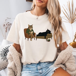 May include: A cream-colored t-shirt featuring a graphic illustration of a tabby cat wearing glasses and a green sweater sitting in a chair, holding a notepad, while a black cat lies on a couch nearby with a small tissue box on the floor.