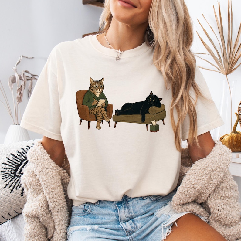 May include: A cream-colored t-shirt featuring a graphic illustration of a tabby cat wearing glasses and a green sweater sitting in a chair, holding a notepad, while a black cat lies on a couch nearby with a small tissue box on the floor.