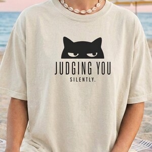 May include: Cream-colored t-shirt featuring a black cat graphic above the text "JUDGING YOU SILENTLY." The shirt has short sleeves and a crew neck. The design is centered on the front of the shirt.