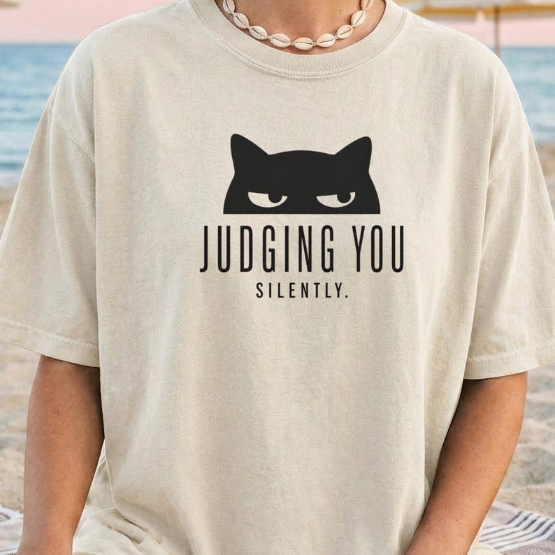 May include: Cream-colored t-shirt featuring a black cat graphic above the text "JUDGING YOU SILENTLY." The shirt has short sleeves and a crew neck. The design is centered on the front of the shirt.