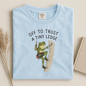 May include: A light blue t-shirt featuring a graphic of a green frog rock climbing on a beige cliff face. The frog wears climbing gear and shoes. Above the frog, the text reads: OFF TO TRUST A TINY LEDGE. The shirt has a Comfort Colors brand label.