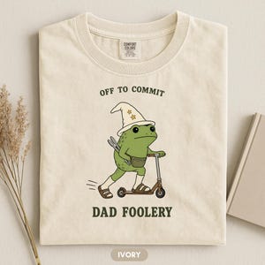 May include: An ivory t-shirt featuring a graphic of a green frog wearing a wizard hat, socks, and sandals while riding a scooter. The frog carries a fanny pack and tongs. The text reads OFF TO COMMIT DAD FOOLERY in dark green lettering.
