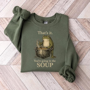May include: An olive green crewneck sweatshirt featuring a graphic of a frog stirring a large metal pot. Above the frog, the text reads "That's it." and below the pot, the text reads "You're going in the SOUP" in a bold, capitalized font.
