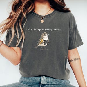 May include: A charcoal gray short-sleeve t-shirt featuring a graphic illustration of a small bird wearing binoculars around its neck. The text "this is my birding shirt" is printed in white, typewriter-style font above the bird illustration.