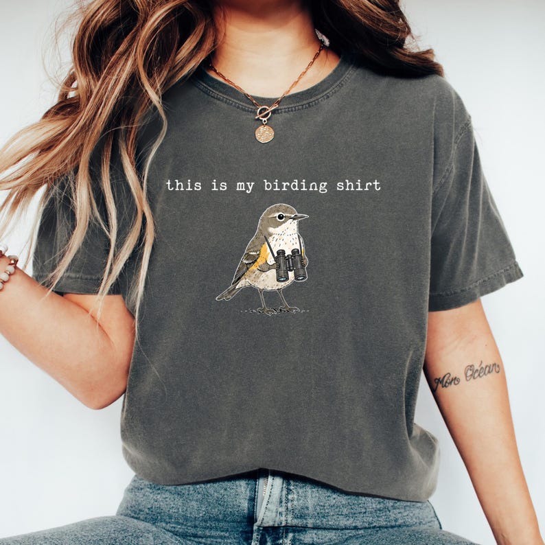 May include: A charcoal gray short-sleeve t-shirt featuring a graphic illustration of a small bird wearing binoculars around its neck. The text "this is my birding shirt" is printed in white, typewriter-style font above the bird illustration.