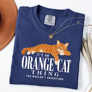 Puede incluir: Una camiseta azul marino con el gráfico de un gato atigrado naranja recostado. Debajo del gato, un texto en blanco dice: IT'S AN ORANGE CAT THING YOU WOULDN'T UNDERSTAND. La camiseta tiene una etiqueta Comfort Colors en el cuello redondo.