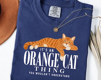 May include: A navy blue t-shirt featuring a graphic of a reclining orange tabby cat. Below the cat, white text reads: IT'S AN ORANGE CAT THING YOU WOULDN'T UNDERSTAND. The shirt has a Comfort Colors label at the neck and a crew neckline design.