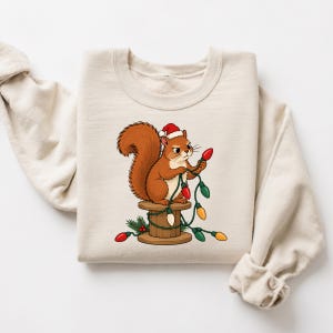 May include: A beige crewneck sweatshirt featuring a graphic of a brown squirrel wearing a red and white Santa hat. The squirrel sits on a wooden spool, tangled in a string of colorful red, green, and yellow Christmas light bulbs.