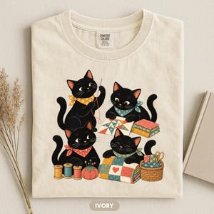 Może przedstawiać: T-shirt w kolorze kości słoniowej z grafiką przedstawiającą cztery czarne koty zajmujące się szyciem i pikowaniem. Koty noszą kolorowe bandany i są otoczone przyborami do szycia, w tym szpulami nici, poduszką na szpilki i koszykiem.
