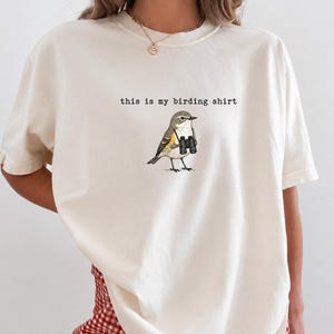May include: An off-white short-sleeve t-shirt featuring a graphic of a small bird holding black binoculars. Above the bird, the text reads "this is my birding shirt" in a typewriter-style font. The shirt has a crew neckline and a relaxed fit.