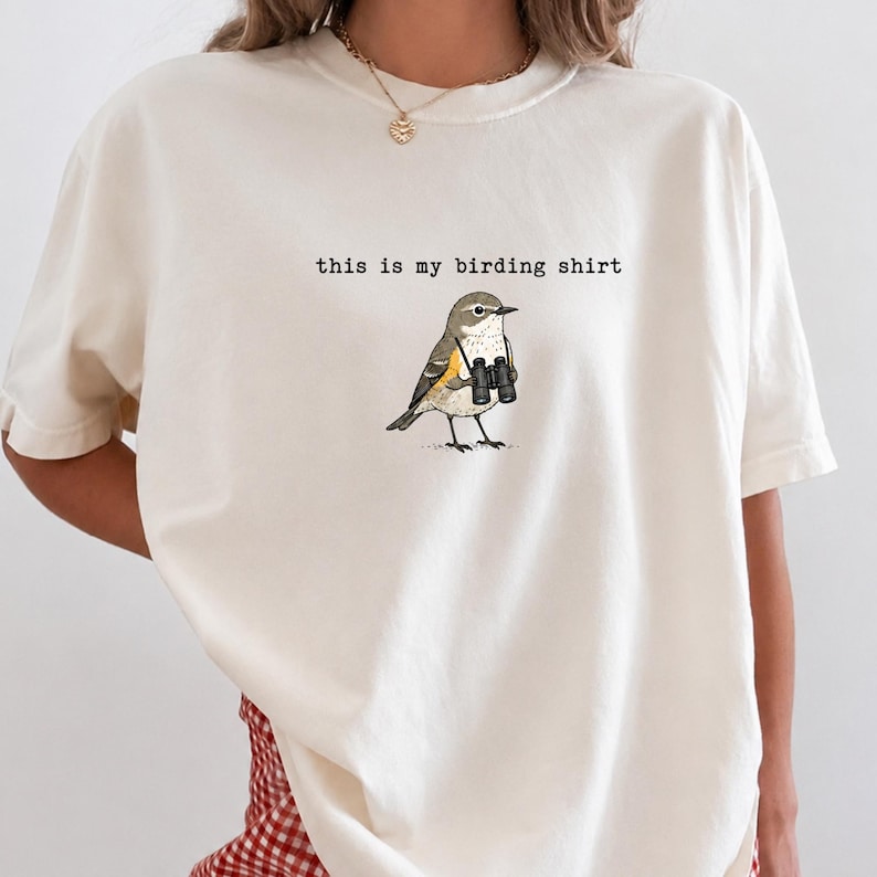 May include: An off-white short-sleeve t-shirt featuring a graphic of a small bird holding black binoculars. Above the bird, the text reads "this is my birding shirt" in a typewriter-style font. The shirt has a crew neckline and a relaxed fit.
