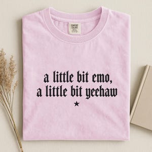 May include: A light pink short-sleeved t-shirt with a crew neckline. The center of the shirt features black text that reads, a little bit emo, a little bit yeehaw, with a small black star symbol centered directly below the text.