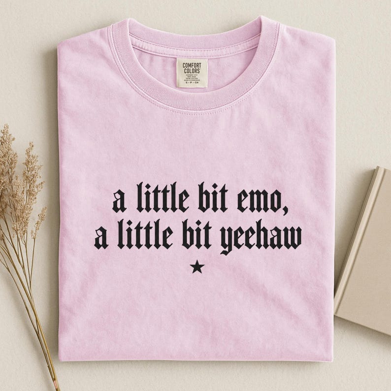 May include: A light pink short-sleeved t-shirt with a crew neckline. The center of the shirt features black text that reads, a little bit emo, a little bit yeehaw, with a small black star symbol centered directly below the text.