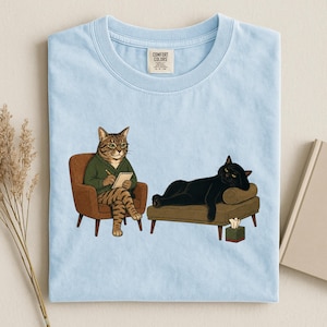 May include: A light blue t-shirt featuring a graphic of a tabby cat in a green sweater sitting in a brown armchair writing on a notepad, while a black cat lies on a brown chaise lounge next to a tissue box. The shirt has a Comfort Colors label.