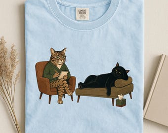 May include: A light blue t-shirt featuring a graphic of a tabby cat in a green sweater sitting in a brown armchair writing on a notepad, while a black cat lies on a brown chaise lounge next to a tissue box. The shirt has a Comfort Colors label.