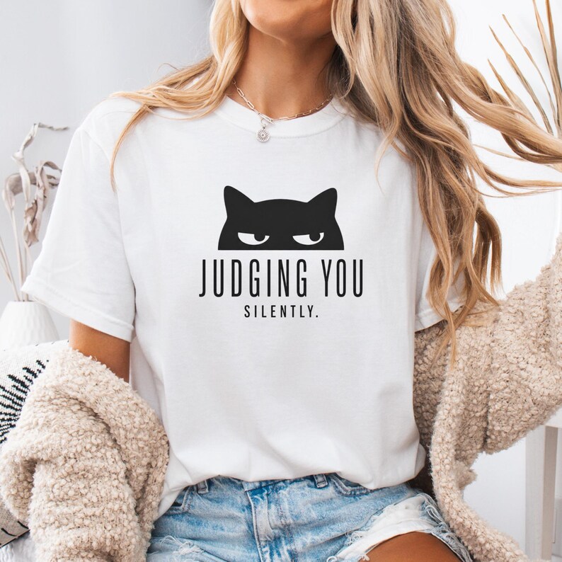 May include: White t-shirt with a black cat graphic above the words "JUDGING YOU SILENTLY." The shirt is a casual style, suitable for everyday wear.