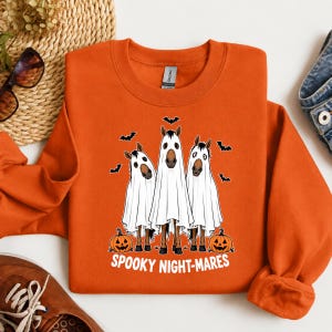 May include: An orange crewneck sweatshirt featuring a graphic of three horses wearing white ghost costumes. The design includes black bats, two orange jack-o'-lantern pumpkins, and the white text "SPOOKY NIGHT-MARES" printed at the bottom.