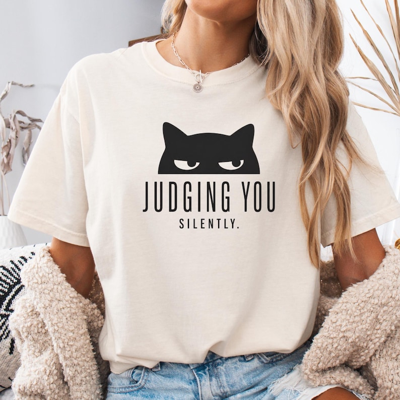 May include: Cream-colored t-shirt with a black cat graphic above the words "JUDGING YOU SILENTLY." The shirt has short sleeves and a relaxed fit. The text and cat graphic are centered on the front of the shirt.