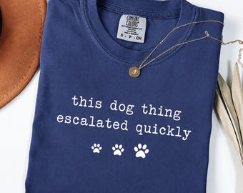 May include: A navy blue crew neck t-shirt featuring the white text "this dog thing escalated quickly" followed by three white paw prints. The shirt has a Comfort Colors brand label inside the collar indicating size S, P, CH and 100% cotton material.