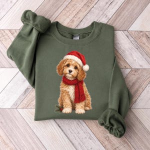 Può includere: Una felpa girocollo verde oliva con la grafica di un cucciolo marrone dorato che indossa una sciarpa di lana rossa e un cappello da Babbo Natale rosso con un pon pon bianco. Il cucciolo è seduto accanto a un rametto di pino con bacche rosse.