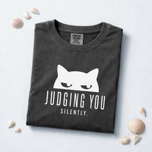 May include: Dark gray t-shirt with a white cat graphic and the words "JUDGING YOU SILENTLY." The shirt is folded and surrounded by seashells. The Comfort Colors tag is visible at the neckline.