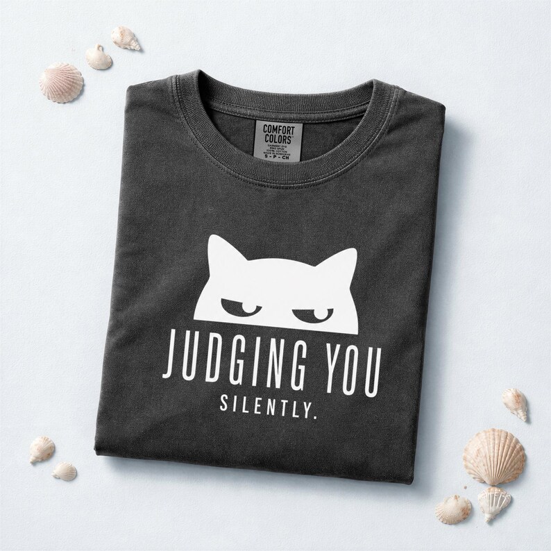 May include: Dark gray t-shirt with a white cat graphic and the words "JUDGING YOU SILENTLY." The shirt is folded and surrounded by seashells. The Comfort Colors tag is visible at the neckline.