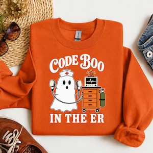 May include: An orange crewneck sweatshirt featuring a graphic of a white ghost wearing a nurse cap and holding defibrillator paddles. Next to the ghost is a medical cart with a monitor and oxygen tank. The text reads CODE BOO IN THE ER.