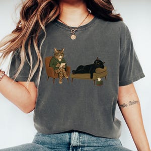 May include: A charcoal gray t-shirt featuring a graphic illustration of a tabby cat wearing a green sweater and glasses, sitting in a brown armchair with a notepad, while a black cat lies on a brown couch next to a small tissue box.