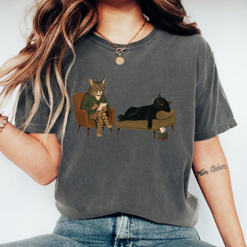 May include: A charcoal gray t-shirt featuring a graphic illustration of a tabby cat wearing a green sweater and glasses, sitting in a brown armchair with a notepad, while a black cat lies on a brown couch next to a small tissue box.