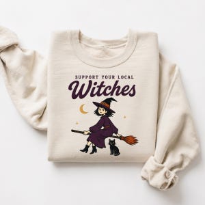 May include: A cream-colored crewneck sweatshirt featuring a graphic print with the text "SUPPORT YOUR LOCAL Witches" in dark purple. The illustration depicts a character in a purple dress and black hat riding a broomstick with a black cat.