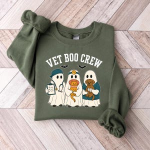 May include: An olive green crewneck sweatshirt featuring a graphic of three ghosts dressed as veterinary staff. The text VET BOO CREW is printed above the ghosts, who are holding a clipboard, an orange cat in a cone, and a brown puppy.