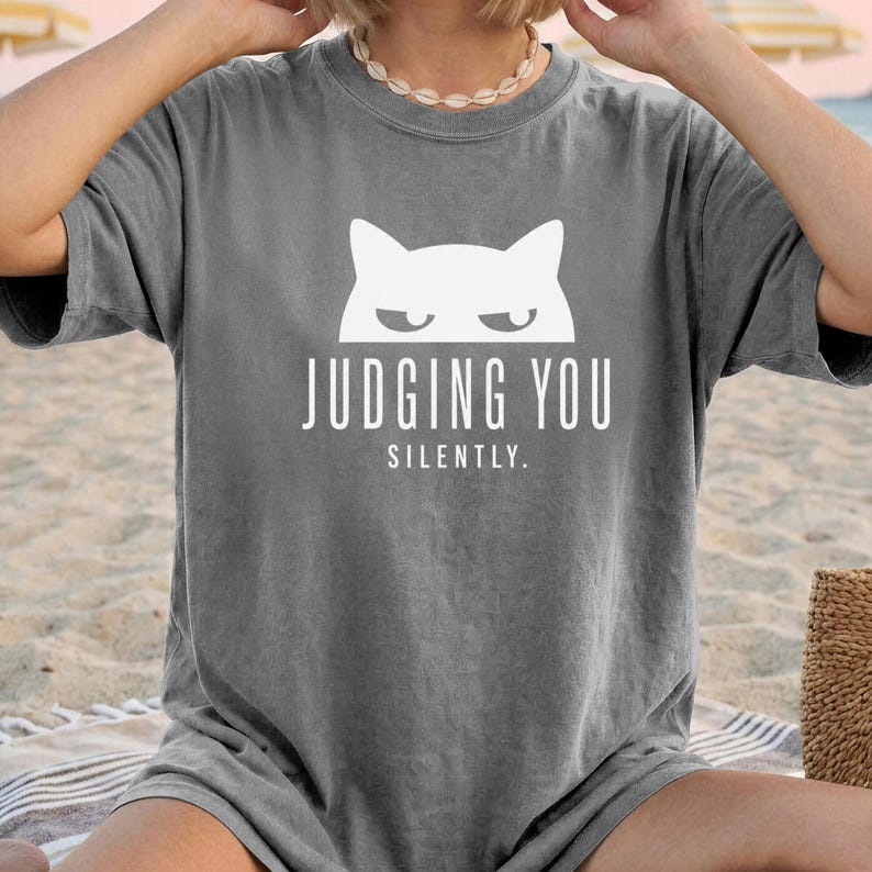 May include: Gray t-shirt with a white cat graphic and the text "JUDGING YOU SILENTLY." The shirt has short sleeves and a relaxed fit. The cat graphic features a minimalist design with a stern expression.