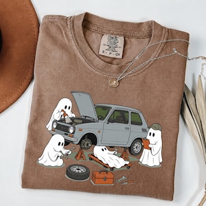 Könnte beinhalten: Ein braunes T-Shirt mit einer Grafik von vier weißen Geistern, die ein graues Auto reparieren. Ein Geist hält eine Taschenlampe unter die geöffnete Motorhaube, ein anderer benutzt einen Schraubenschlüssel, einer liegt auf einem Rollbrett.