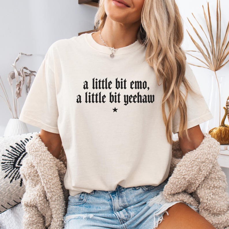 May include: A cream-colored short-sleeve t-shirt featuring black text that reads a little bit emo, a little bit yeehaw with a small star symbol centered below the text. The shirt has a crew neckline and a relaxed fit, worn with blue denim shorts.