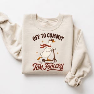 Op de afbeelding: Een beige sweater met ronde hals en een afbeelding van een witte gans met een rode bril en een rode sjaal op een step. De tekst erboven luidt OFF TO COMMIT en de tekst eronder luidt TOM FOOLERY in donkerbruine letters.