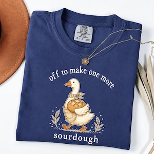 Op de afbeelding: Een marineblauw t-shirt met korte mouwen en een afbeelding van een witte gans met een geruite halsdoek en schort, die een brood draagt. De tekst "off to make one more sourdough" staat in witte letters op het shirt. Het heeft een ronde hals.
