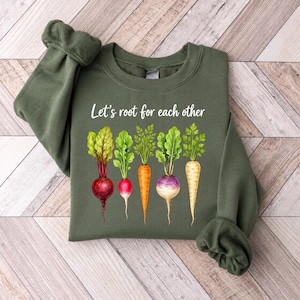 May include: An olive green crewneck sweatshirt featuring a graphic of five colourful root vegetables including a beet, radish, carrot, turnip, and parsnip. The white text above the vegetables reads, Let's root for each other, in a script font.