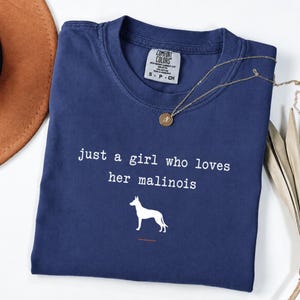 Könnte beinhalten: Ein marineblaues Kurzarm-T-Shirt mit dem weißen Text "just a girl who loves her malinois" über einer weißen Hundesilhouette. Das T-Shirt hat ein Comfort Colors Markenetikett am inneren Halsausschnitt und eine dünne goldfarbene Kettenhalskette.